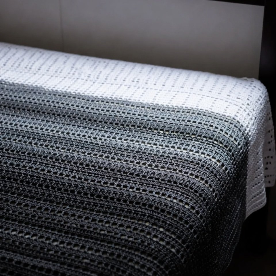 White to charcoal grayscale filet crochet bed sheet with airy mesh pattern, laid flat on a minimalist king bed.